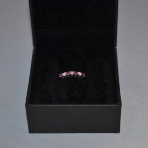 May include: A white gold band ring with pink and white gemstones. The ring is in a black velvet ring box.