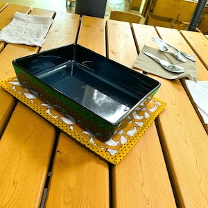 May include: A rectangular, dark blue serving dish with a green trim, on a yellow placemat patterned with white chickens. The dish sits on a wooden table, with cutlery and napkins in the background.