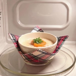 May include: A cream-coloured ceramic bowl containing oatmeal topped with orange fruit slices and green herbs, resting inside a fabric bowl cosy featuring a red, green, and black plaid pattern, all placed on a glass microwave turntable.