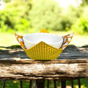 May include: A white ceramic bowl sits within a yellow fabric bowl cosy. The cosy features a black polka dot pattern on the base and a white and orange leaf pattern on the sides. It rests on a wooden surface.