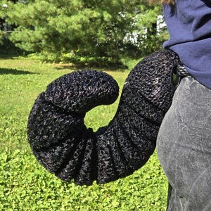 May include: A black, segmented, curled tail accessory featuring a textured, mesh-like surface. The tail is attached to a circular ring at the base, which is secured to the waist area of a pair of grey pants, creating a curved, coiled shape.