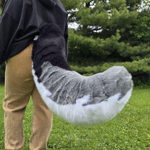 May include: A long, plush faux fur tail featuring a gradient color pattern that transitions from black at the base to shades of grey in the middle and ends with a solid white tip. The tail has a thick, curved shape and a soft, textured appearance.