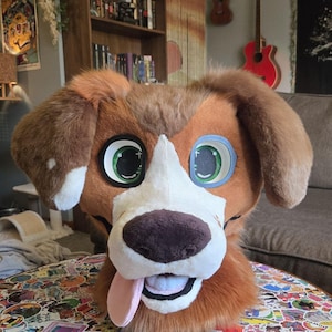 Könnte beinhalten: Ein braun-weißer Hunde-Fursuit-Kopf mit großen grünen Augen mit schwarzen Pupillen, einer dunkelbraunen Nase und einer rosa Zunge, die aus dem Mund hängt. Der Kopf ist mit weichem, kurzflorigem Kunstfell bedeckt und hat Schlappohren.