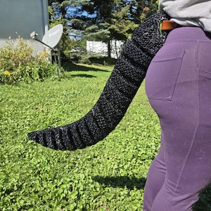 May include: A long, segmented black tail accessory with a textured, crocheted appearance. The tail features a brown leather-like loop attachment with a silver-toned metal ring, designed to be worn at the waist over purple leggings and a grey shirt.
