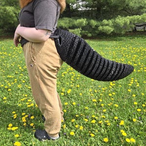 May include: A black and gray striped tail prop, attached to a person wearing tan pants and a gray shirt. The tail is a furry accessory, likely for a costume. The background is a field of green grass and yellow flowers.