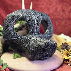 May include: A dark gray, textured mask with large eye openings and a prominent snout. Two small, white antler-like protrusions extend from the top. The mask rests on a pinkish-tan surface against a red backdrop.