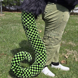 May include: A long, plush tail featuring a black and neon green checkered pattern. The base of the tail is covered in thick, solid black faux fur, while the main length displays a repeating square grid design in alternating black and green colors.