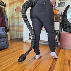 Può includere: Un accessorio a forma di coda nero e strutturato, forse per un costume, si curva dalla schiena di una persona che indossa pantaloni neri. La coda è a strisce e si assottiglia fino a un'estremità arrotondata. Un secondo accessorio simile è visibile.