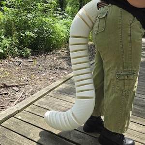 May include: A long, segmented, off-white tail accessory with a textured, foam-like appearance. The tail features a series of horizontal, parallel ridges along its length, creating a flexible, articulated structure that curves downward from the waist.