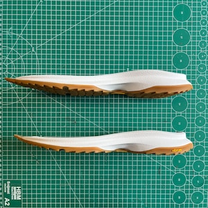 May include: Two white and brown shoe insoles with a textured design and a Vibram logo. The insoles are designed for athletic footwear and are shown on a green cutting mat.