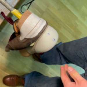 以下が含まれることがあります： 白い義足に茶色の靴が乗っているクローズアップ。義足はストラップで人の足に固定されています。義足には「14」という番号が書かれています。
