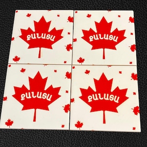 Puede incluir: Cuatro posavasos blancos con diseños de hojas de arce rojas y la palabra "PULUSU" impresa en cada uno. Los posavasos están sobre una superficie negra.