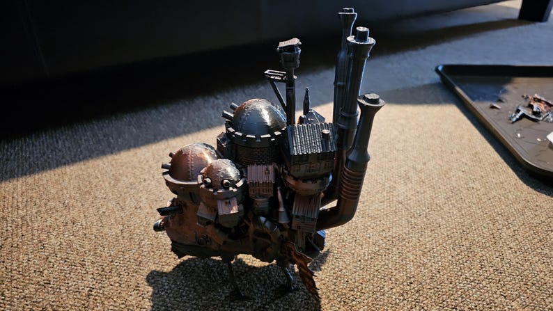 Op de afbeelding: Een gedetailleerd model van een vliegend huis met meerdere schoorstenen en een rond dak. Het huis is gemaakt van donkergrijs en bruin plastic en heeft een steampunk-esthetiek.