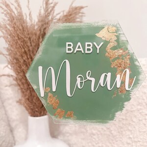 May include: A green hexagonal sign with gold accents and the text "BABY Moran" in white lettering. The sign is attached to a clear plastic stick.