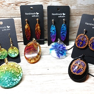 May include: Assortment of handmade resin jewelry featuring colorful pendants and earrings. The pendants are round and oval shaped with abstract designs. The earrings are oval shaped with floral patterns. The jewelry is displayed on black cards with the text "Handmade with love".