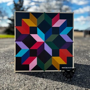 May include: A colorful geometric barn quilt with a starburst design. The quilt is painted on a wooden board with a black background and features shades of red, pink, yellow, green, blue, and purple. The text "BARN QUILTS BY KARLY" is printed in white at the bottom of the board.
