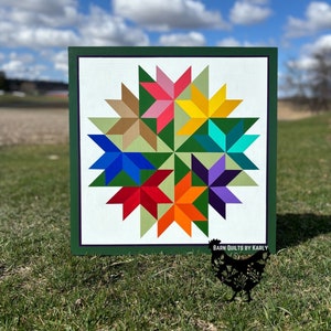May include: A green framed barn quilt with a white background and a colorful starburst design. The quilt features eight points with alternating colors of green, yellow, orange, red, blue, purple, teal, and pink. The quilt is signed "Barn Quilts by Karly" in the bottom right corner.