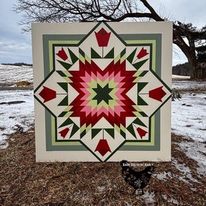 May include: A large, square barn quilt with a starburst design. The quilt features a cream-colored background with a central star pattern in shades of red, pink, green, and dark green. Red tulip shapes are placed around the star. The text "BARN QUILTS BY KARLY" is visible.