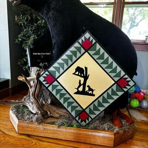 May include: A black bear taxidermy mount with a decorative square sign featuring a bear and dogs silhouette. The sign has a cream-colored center with a black border and green and red accents. The base is a wooden box.