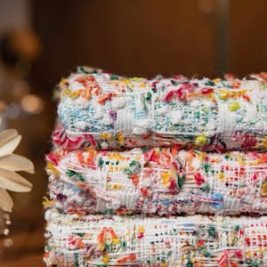 Può includere: Una pila di quattro rotoli di tessuto tweed colorato. Il tessuto è bianco con macchie di blu, rosso, giallo e verde. I bordi del tessuto sono sfilacciati. I rotoli sono impilati su una superficie di legno.