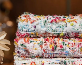 May include: Four rolls of colourful tweed fabric are stacked. The fabric is white with flecks of blue, red, yellow, and green. The edges are fringed. The rolls are on a wooden surface. The fabric is suitable for clothing.