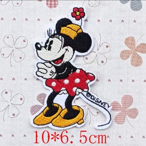 Peut inclure: Patch thermocollant brodé représentant un dessin animé de Minnie Mouse portant une jupe rouge à pois blancs et des chaussures jaunes. Le patch mesure 10 x 6,5 cm et comprend le texte "©DISNEY".