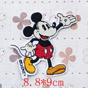 Könnte beinhalten: Ein gestickter Bügel-Patch mit Mickey Mouse in einem roten und schwarzen Outfit, der mit einem breiten Lächeln winkt. Der Patch ist 8,8 x 9 Zentimeter groß und enthält das Copyright-Symbol und den Text "DISNEY".