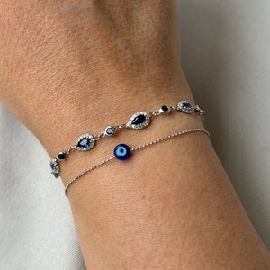 May include: Two silver bracelets. The top bracelet features teardrop-shaped blue gemstones with surrounding clear stones. The bottom bracelet has a single blue evil eye charm. Both bracelets are on a wrist.