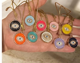 Puede incluir: Una colección de colgantes redondos y coloridos con un diseño de ojo. Cada colgante tiene un fondo de color diferente, incluyendo negro, naranja, azul, rosa y amarillo. Los colgantes están unidos a ganchos de metal dorado.