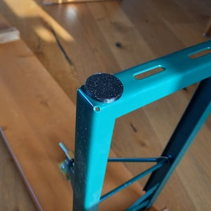May include: Close-up of a teal metal table leg with black rubber feet. The leg is attached to a wooden table top.