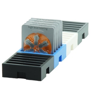 May include: A blue, white and black plastic organiser with compartments for storing hearing aid batteries. The organiser has a label that reads "high level hearing".