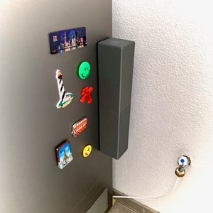 Può includere: Un frigorifero grigio scuro con diversi magneti souvenir colorati. Una maniglia alta e rettangolare, grigio scuro, è montata sul lato. I magneti includono un paesaggio urbano, un faro, un autobus rosso e una faccina sorridente.
