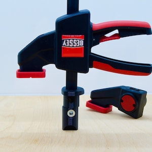 May include: A black and red quick-release clamp with the word "BESSEY" in white on a red rectangle. The clamp is on a light brown wooden surface.