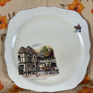May include: A white plate with a wavy edge, featuring a detailed illustration of a historic building and figures. The plate has a gold trim and the text "Old Coach House, Stratford". The scene depicts people and horses.