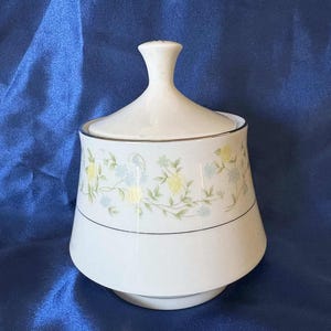 May include: A white ceramic sugar bowl with a lid, featuring a delicate floral pattern in yellow and blue. The bowl has a gold trim and a pedestal base. The lid has a small handle.