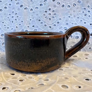 May include: A ceramic mug with a wide, low-profile cylindrical body and a single curved handle. The exterior features a dark brown glaze with a mottled, textured finish that transitions into a lighter, speckled bronze tone near the base.