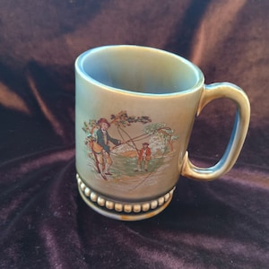 Puede incluir: Una taza de cerámica verde y marrón con una escena de dos hombres pescando. La taza tiene un borde con cuentas y un asa.