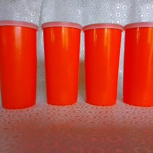 May include: Four orange plastic cups with white lids. The cups are lined up in a row on a white surface with a patterned design.