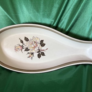 May include: A white ceramic spoon rest with a floral rose design. The spoon rest has a light brown border and a curved shape. The floral design features white and pink roses with green leaves.
