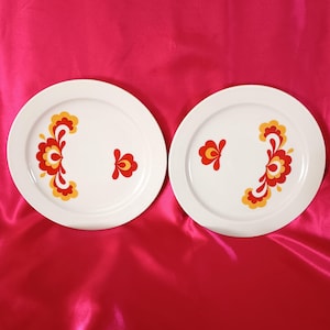 May include: Two white ceramic plates with a red, yellow, and white floral design. The plates are on a pink background.