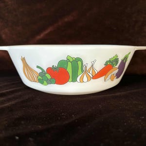 May include: White ceramic bowl with a colorful vegetable design. The bowl features illustrations of an onion, tomato, broccoli, bell pepper, garlic, carrot, mushroom, and eggplant. The bowl has two small handles on either side.