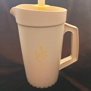 Può includere: Un'ampolla di plastica bianca con un coperchio giallo e un motivo floreale giallo. L'ampolla ha un manico e un beccuccio.