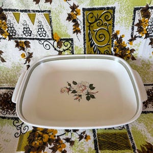 Puede incluir: Una fuente de horno rectangular de cerámica blanca con esquinas redondeadas y asas laterales integradas. El centro de la fuente presenta una ilustración floral detallada de una rosa blanca con hojas verdes y pequeños capullos.