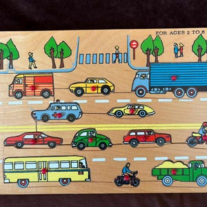 May include: A wooden peg puzzle featuring a colorful street scene with various vehicles including a bus, truck, cars, and motorcycles. The board displays the text FOR AGES 2 TO 6 and includes illustrations of trees, a crosswalk, and people.