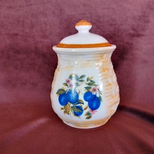 May include: A white ceramic jar with a lid, featuring a woven pattern and a floral design with blue plums and pink flowers.