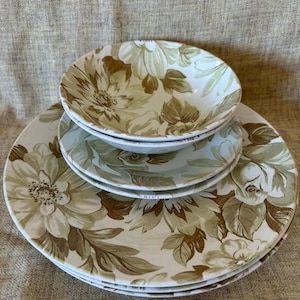 May include: A set of four white ceramic plates and bowls featuring a floral design in brown and green hues. The plates and bowls are arranged in a stack, with the largest plate at the base and the smallest bowl at the top.