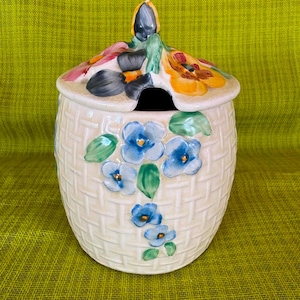 May include: A cream-coloured ceramic jar with a basket-weave texture, featuring hand-painted blue flowers on the body and a lid adorned with colourful pansies. The lid includes a small cutout for a spoon and a central handle shaped like a bud.