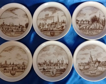 May include: Six white ceramic coasters with brown illustrations of Dutch cityscapes. Each coaster features a different city with its name and coat of arms printed on the bottom.