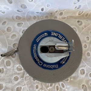 Puede incluir: Un flexómetro gris Chesterman Rabone Everline con una etiqueta azul y blanca que dice "Made in England". El flexómetro tiene un asa de metal plateado. El fondo es blanco.