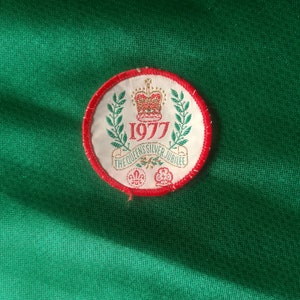 Może przedstawiać: Okrągła naszywka w kolorze czerwonym, białym i zielonym z koroną i tekstem "1977 The Queen's Silver Jubilee".